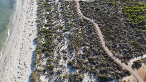 Drone-aerial-following-a-running-track-by-a-white-sand-pristine-beach-with-greenery