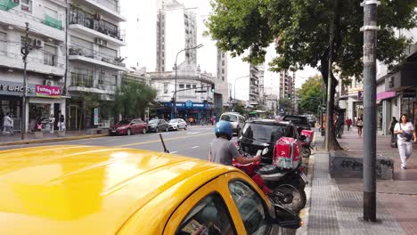 El-Conductor-De-La-Moto-De-Entrega-Espera-Fuera-Del-área-Comercial-En-El-Tráfico-De-La-Concurrida-Avenida-Latina-Pedidosya,-Taxi-Amarillo-Negro-Del-área-Metropolitana-De-La-Ciudad-De-Buenos-Aires