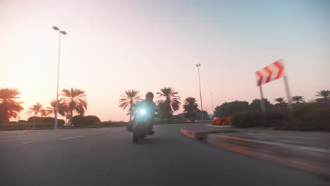 Eine-Rollaufnahme-Eines-Schwarzen-Motorrads,-Das-In-Einer-Verfolgungsjagd-Um-Einen-Kreisverkehr-Kreist,-Ein-Schwarz-Gekleideter-Motorradfahrer,-Der-Während-Eines-Epischen-Sonnenuntergangs-Durch-Abu-Dhabi-Fährt