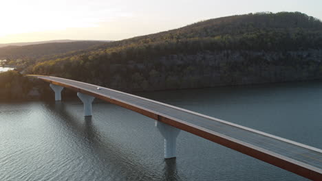 Static-aerial-footage-of-a-car-driving-across-a-bridge-with-a-cliff-and-forest-in-the-background-during-golden-hour-in-Tennessee