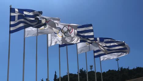 Olympische-Und-Griechische-Flaggen-Schweben-In-Zeitlupe-Im-Wind-Mit-Einem-Blauen-Himmel-Im-Hintergrund