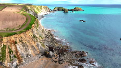 Drone-flying-over-headland-with-sandy-cliffs-eroding-into-the-sea-and-rockfalls-on-the-beach-with-crystal-clear-water-and-deep-blue-colours-Copper-Coast-in-Waterford-Ireland-on-a-spring-afternoon