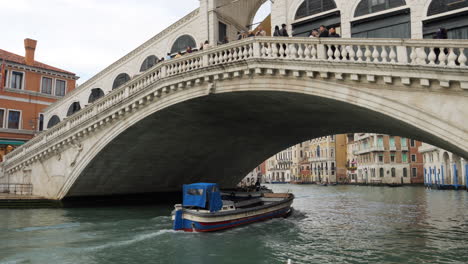 Boat-passing-under-Rialto-Bridge,-tourists-taking-pictures,-Venice,-Italy