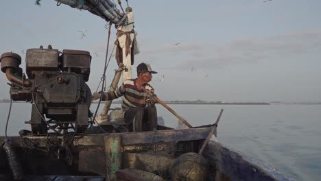 Un-Pescador-Tradicional-Lanza-Hábilmente-Su-Red-De-Pesca-Usando-Bambú-Desde-Su-Viejo-Barco-Impulsado-Por-Un-Viejo-Motor,-Mientras-Las-Gaviotas-Vuelan-Con-Gracia,-Creando-Una-Hermosa-Escena.