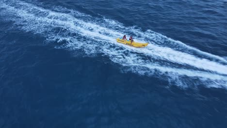 Couple-Riding-Banana-Sled-Tube-on-Sea-Water-Tugged-By-Jet-Ski,-Drone-Follow-Shot