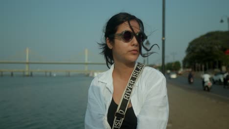 Young-Indian-woman-putting-on-her-sunglasses-next-to-a-promenade-and-a-river