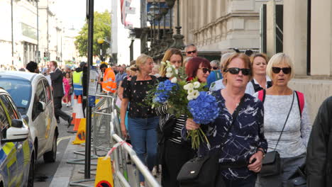 Scene-of-remembrance-as-people-carrying-flowers,-walk-the-barricaded-walkway,-after-Queen-Elizabeth-II's-funeral,-with-the-British-flag-hanging-in-the-backdrop