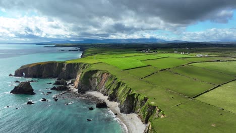 Drohnenaufnahme-Einer-Atemberaubenden-Küste-Mit-Smaragdgrünen-Feldern-Und-Bergen-Im-Hintergrund,-Die-Dramatische-Landschaft-Von-Waterford,-Irland-Im-Frühling