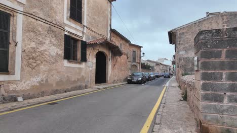 Walking-the-street-in-town-Algaida,-Balearic-island-of-Mallorca-with-limestone-houses-street-parked-cars-in-April
