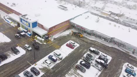 Aerial-view-over-snowplough-clearing-snow-from-a-shopping-centre-car-park,-Montreal-Canada
