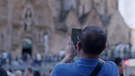 Man-capturing-Sagrada-Familia-on-smartphone,-Barcelona,-tourists-around,-day,-rear-view,-blurred-background