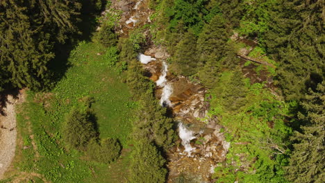 Aerial-of-a-small-mountain-creek-surrounded-by-a-green-forest-and-meadow