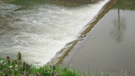 Rushing-waters-at-a-river-weir,-creating-a-white-frothy-texture,-reflecting-surrounding-nature