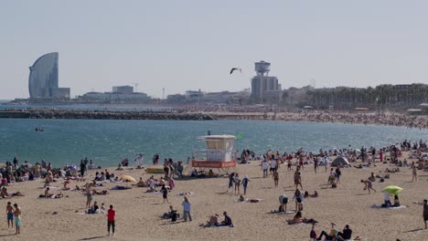 überfüllter-Strand-Von-Barcelona-An-Einem-Sonnigen-Tag,-Skyline-Der-Stadt-Im-Hintergrund,-Blauer-Himmel