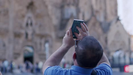 Hombre-Con-Camisa-Azul-Capturando-La-Sagrada-Familia-Con-Un-Teléfono-Inteligente,-Turistas-En-El-Fondo,-Durante-El-Día,-Barcelona