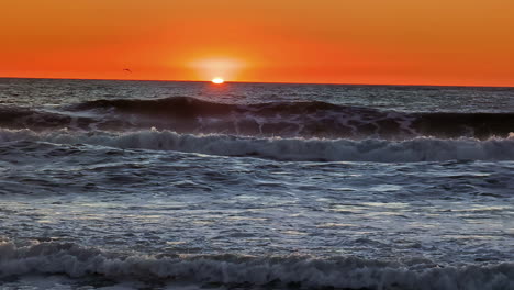 Timelapse-shot-of-majestic-sun-setting-along-orange-sky-over-the-horizon-with-waves-crashing-along-the-sandy-beach-during-evening-time