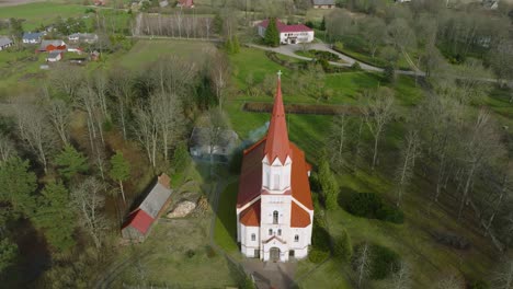 Aerial-view-of-a-white-church-with-red-roof-on-a-sunny-spring-day,-wide-angle-drone-orbit-shot