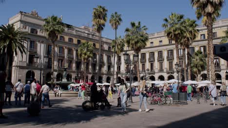 Sunny-day-at-a-bustling-Barcelona-plaza-with-people-and-palm-trees,-wide-shot