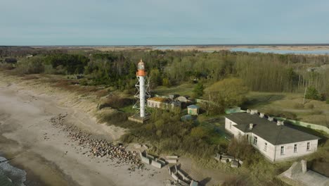Aerial-establishing-view-of-white-colored-Pape-lighthouse,-Baltic-sea-coastline,-Latvia,-white-sand-beach,-large-waves-crashing,-sunny-day-with-clouds,-wide-drone-shot-moving-forward