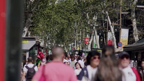 Geschäftige-Straßenszene-In-Barcelona-Mit-Fußgängern,-Bäumen-Und-Ladenschildern,-Tageslicht,-Unscharfer-Vordergrund