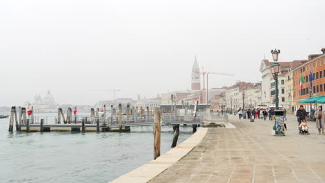 Gente-Caminando-Frente-Al-Mar-A-Lo-Largo-De-La-Laguna-Veneciana-Con-El-Campanario-De-San-Marcos-En-La-Distancia-En-Venecia,-Italia