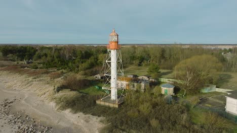 Aerial-establishing-view-of-white-colored-Pape-lighthouse,-Baltic-sea-coastline,-Latvia,-white-sand-beach,-large-waves-crashing,-sunny-day-with-clouds,-wide-drone-orbit-shot