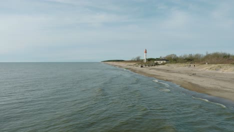 Aerial-establishing-view-of-white-colored-Pape-lighthouse,-Baltic-sea-coastline,-Latvia,-white-sand-beach,-large-waves-crashing,-sunny-day-with-clouds,-wide-drone-shot-moving-froward-ascending