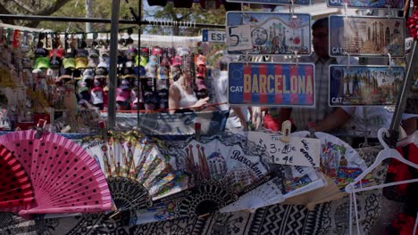 Puesto-De-Souvenirs-Colorido-En-Barcelona-Con-Fans-Y-Postales,-Ambiente-De-Mercado-Bullicioso,-Durante-El-Día