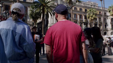 Man-holding-picture-among-crowd-in-sunny-Barcelona-square,-casual-urban-life