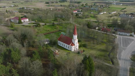 Aerial-view-of-a-white-church-with-red-roof-on-a-sunny-spring-day,-distant-wide-angle-drone-orbit-shot