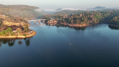 Drone-advancing-over-Khwae-Noi-Lake,-revealing-the-Songkalia-Bridge-in-the-background,-in-Songklaburi,-Thailand