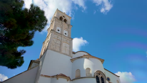 Low-angle-view-of-a-white-church-with-a-stone-bell-tower-against-a-blue-sky-with-fluffy-clouds,-surrounded-by-greenery,-located-in-Lefkara,-Cyprus