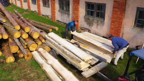 Two-workers-stacking-lumber-in-pile-outdoors-next-to-sawmill-for-drying-in-sun