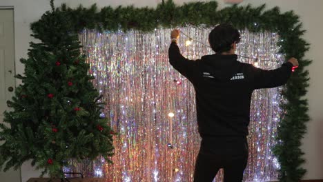 Young-man-putting-up-Christmas-lights-decoration-in-studio-set-with-Christmas-tree