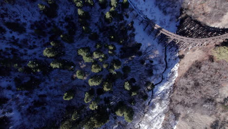 Aerial-top-down-of-a-snow-covered-valley-in-a-pine-forest-in-the-mountains,-old-narrow-gauge-railway-tracks-in-the-foreground