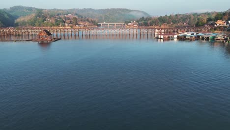 Un-Dron-Deslizándose-Sobre-El-Río-Khwae-Noi,-Acercándose-Al-Puente-Mon-En-Songklaburi,-Tailandia,-Capturando-La-Tranquila-Belleza-Del-Entorno