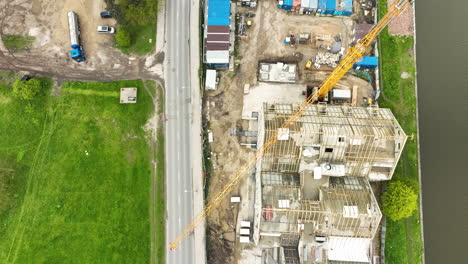 Aerial-view-of-a-construction-site-with-a-crane-next-to-a-road,-flanked-by-a-green-field-and-industrial-containers