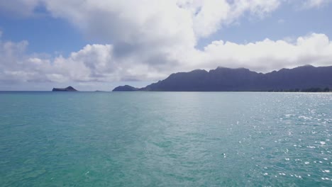 drone-footage-across-the-turquoise-water-toward-silhouetted-mountains-on-the-island-of-Oahu-in-the-Hawaiian-islands-white-clouds-and-blue-sky
