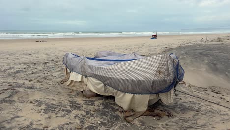 Boat-sheltered-by-a-tarp-and-a-net-on-a-deserted-beach-on-a-gray-day