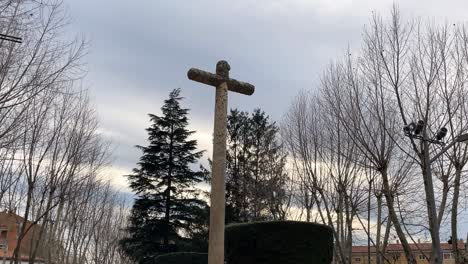 Rodaje-De-Una-Cruz-De-Piedra-De-Granito-Con-Su-Pedestal-Con-Inscripciones-Ubicada-En-Los-Alrededores-De-La-Iglesia-De-San-Nicolás-Con-El-Fondo-De-Un-Parque-Con-árboles-En-Una-Mañana-De-Invierno