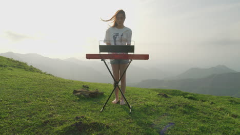 drone-approaching-young-female-musician-performing-playing-piano-keyboard-in-pure-nature-at-sunset-mountains-green-valley-landscape