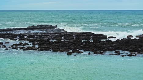 Panorámica-Aérea-De-Afloramientos-Rocosos-Bajos-Mientras-El-Agua-Del-Océano-Salpica-De-Las-Olas-En-Las-Llanuras-De-Basalto.