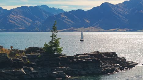 Aerial-trucking-pan-around-rocky-island-reveals-sailboat-calmly-floating-in-middle-of-lake