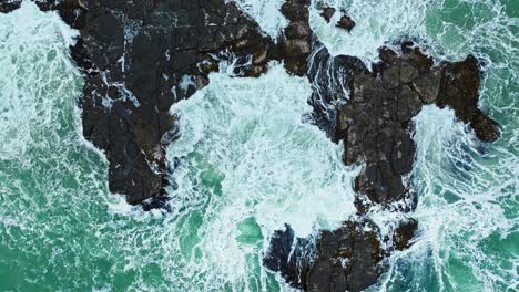 Aerial-top-down-ascends-as-water-pulses-erupting-over-rocky-cliff-edge-in-ocean