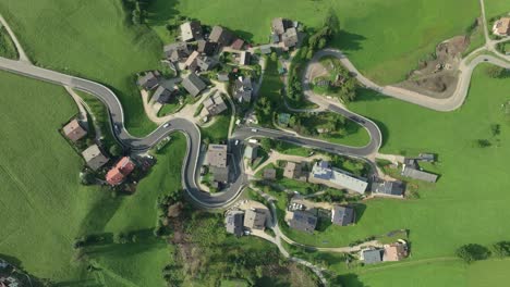 An-overhead-aerial-tilt-down-view-of-cars-moving-on-a-winding-road-of-the-small-village-of-La-Val,-South-Tyrol,-Italy-with-it's-lush-green-steep-grass-covered-hills