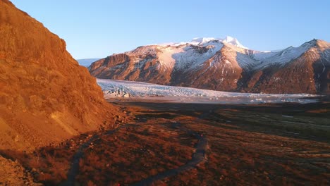 Drone-Estableciendo-Una-Toma-De-Dos-Valientes-Exploradores-Caminando-Por-Un-Sendero-Solitario-Hacia-El-Glaciar-Snaefellsjokull-En-La-Costa-Sur-De-Islandia-En-Un-Paisaje-Bañado-Por-Una-Luz-Dorada-Y-Montañas-Escarpadas-Cubiertas-De-Nieve