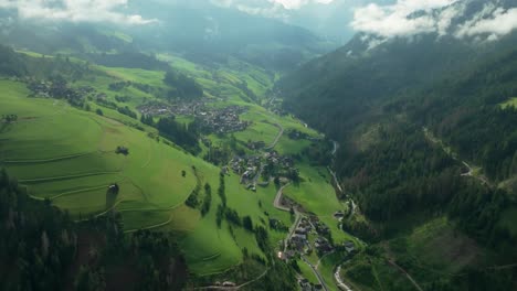 Una-Vista-Aérea-Inclinada-Hacia-Abajo-Del-Valle-Del-Pequeño-Pueblo-De-La-Val,-Tirol-Del-Sur,-Italia,-Con-Sus-Sinuosos-Caminos-Que-Atraviesan-Las-Empinadas-Colinas-Cubiertas-De-Hierba.