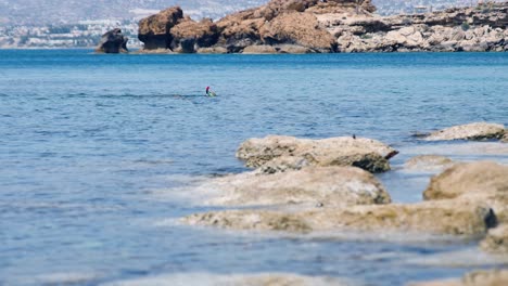 A-person-snorkeling-in-a-blue-bay,-lagoon-on-the-Mediterranean-Sea,-windy-and-sunny-weather,-rocky-shore,-with-mountains-in-the-background,-Cyprus,-Paphos,-at-the-end-of-a-bright-red-snorkel-tube
