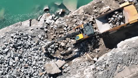 aerial-Top-view-of-an-excavator-loading-stones-into-a-truck-by-a-quarry-lake