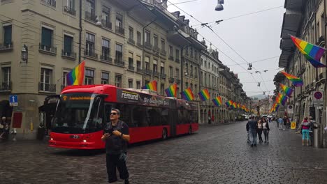 Downtown-Street-in-Bern,-Switzerland-During-LGBT-Pride,-Rainbow-Flags,-Electric-Bus-and-People